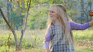 Beautiful long blonde hair girl holds yellow leaves on nature background. Smiling child plays in autumn park sunny day