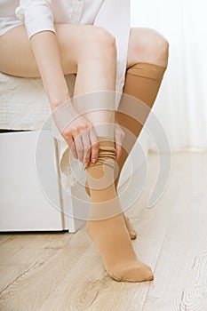 Beautiful long female legs in stockings. Girl putting on stockings at home in a white room. Beige knee socks. Varicose