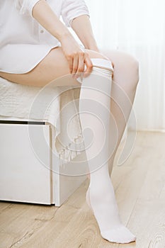 Beautiful long female legs in stockings. Girl putting on stockings at home in a white room. White hosiery. Varicose