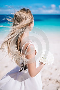 Beautiful long hair bride in long white dress standing on the beach with white bouquet looking to the sea