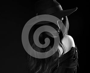 Beautiful long hair woman posing in black shirt and fashion elegant hat in dramatic light on dark shadow background. Closeup art