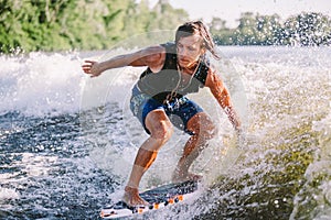 Beautiful long haired blond wakesurf in vest on board along waves of lake. Athletic male athlete wakes surfing in summer on river photo