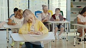 Beautiful long-haired blonde pupil sits at a desk at school and fills out tests, and the teacher in the background help