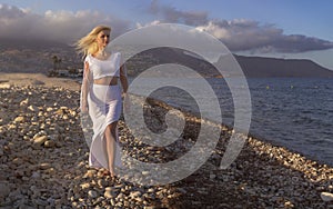 Beautiful long-haired blonde woman walking by the sea at sunset while the wind moves her hair