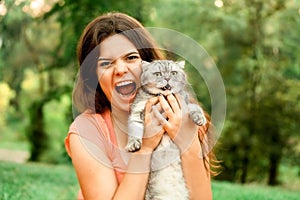 Beautiful long-haired brunette girl in a park sitting with a cat. The girl growls at the cat and bites. Photoshoot with a pet cat