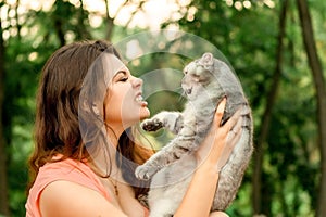 Beautiful long-haired brunette girl in a park sitting with a cat. The girl growls at the cat and bites. Photoshoot with a pet cat