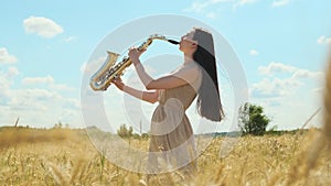 Beautiful long-haired brunette woman, jazz performer plays saxophone outdoors in field with blue sky on background. lady