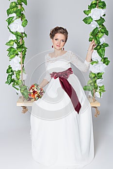 Beautiful lovely bride is sitting on a swing with a beautiful bouquet of colorful flowers in a white dress with evening hairstyle