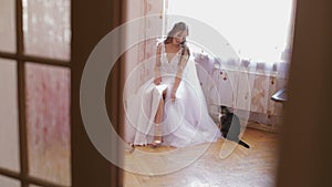 Beautiful and lovely bride in wedding dress sit on a chair and look at her shoes. Cat sits near woman. Wedding morning