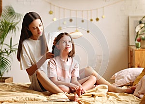 Beautiful lovely older sister helping with hairstyle to younger in the morning while sitting on the bed at home
