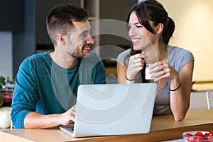 Beautiful lovely young couple using their laptop and having breakfast in the kitchen at home.