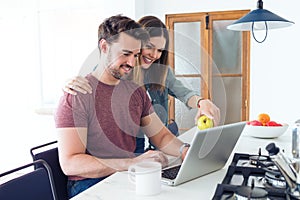 Beautiful lovely young couple using their laptop and having breakfast in the kitchen at home