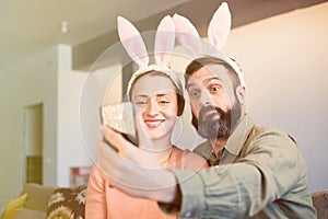 Beautiful loving couple making selfie on smartphone with pink rabbit ears on head. Happy family preparing for Easter.