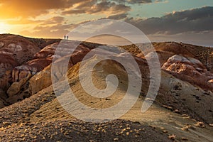 Beautiful low angle shot of two tiny human figures walking towards sunset on top of massive red mountain in Kyzyl-Chin valley,