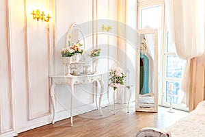 Beautiful luxury classic white bright clean interior bedroom in baroque style with mirror, table, large window, and flower