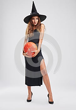 Beautiful, magical witch girl in a hat, holding a pumpkin on a gray background. Halloween concept.