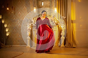 A beautiful majestic solemn fat queen in a red elegant dress in a beautiful hall in the palace. A chubby model poses