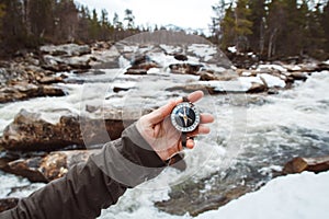 Beautiful male hand holds a magnetic compass against the background of mountain river, rocks and forest. The concept of finding