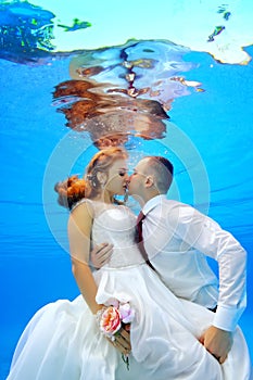 Beautiful man and woman in wedding dresses hugging and kissing underwater in the swimming pool on a background of sun rays