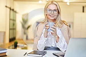 Beautiful mature businesswoman in glasses smiling at camera and drinking coffee while working on the laptop in office