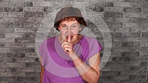 A beautiful mature woman in casual clothes is doing silencing gesture with index finger at her lips on a gray brick