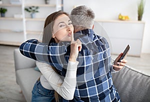 Beautiful mature woman hugging her husband, showing HUSH gesture, having secret affair, texting her lover on smartphone