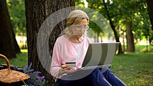 Beautiful mature woman inserting card number on laptop pc in park, shopping