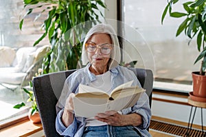 Beautiful mature woman reading book, feeling cozy and happy. Weekend activity for older woman, relaxing at home.