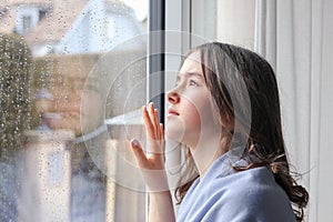 Beautiful melancholic teenager girl in light blue shawl  looking outside through raindrops on wet window