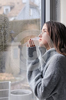 Beautiful melancholic teenager girl in warm grey sweater looking outside through raindrops on wet window at sky at spring rainy da