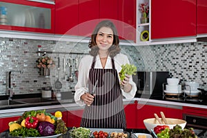 Beautiful middle age Asian woman housewife wearing apron standing new red tone kitchen and holding fresh green vegetable and knife