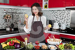 Beautiful middle age Asian woman housewife wearing apron standing new red tone kitchen and holding fresh lemon vegetable
