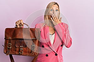 Beautiful middle age blonde woman holding business bag covering mouth with hand, shocked and afraid for mistake