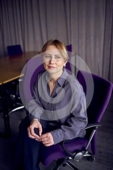 beautiful middle age business woman wearing gray shirt, wide leg pants abd black stilettos sit in purple office chair in modern