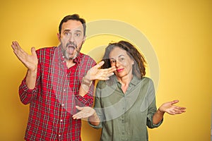 Beautiful middle age couple over isolated yellow background clueless and confused expression with arms and hands raised