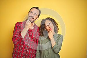 Beautiful middle age couple over isolated yellow background with hand on chin thinking about question, pensive expression