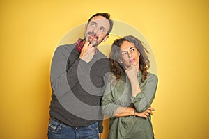 Beautiful middle age couple over isolated yellow background with hand on chin thinking about question, pensive expression