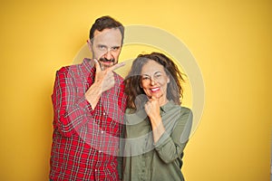 Beautiful middle age couple over isolated yellow background looking confident at the camera with smile with crossed arms and hand