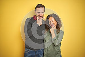 Beautiful middle age couple over isolated yellow background looking confident at the camera with smile with crossed arms and hand