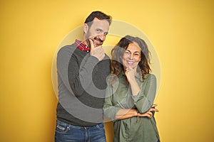 Beautiful middle age couple over isolated yellow background looking confident at the camera smiling with crossed arms and hand