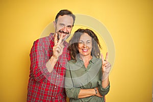 Beautiful middle age couple over isolated yellow background smiling with happy face winking at the camera doing victory sign