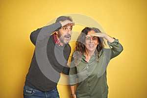 Beautiful middle age couple over isolated yellow background very happy and smiling looking far away with hand over head