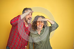 Beautiful middle age couple over isolated yellow background very happy and smiling looking far away with hand over head