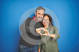 Beautiful middle age couple together standing over isolated blue background smiling in love showing heart symbol and shape with