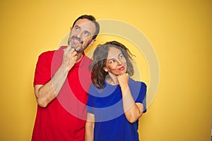 Beautiful middle age couple together standing over isolated yellow background with hand on chin thinking about question, pensive