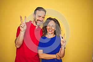 Beautiful middle age couple together standing over isolated yellow background smiling with happy face winking at the camera doing