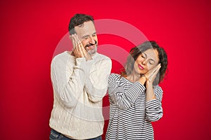 Beautiful middle age couple wearing winter sweater over isolated red background sleeping tired dreaming and posing with hands