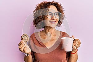 Beautiful middle age mature woman drinking a cup of coffee and cookie smiling looking to the side and staring away thinking