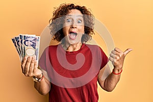 Beautiful middle age mature woman holding 5000 japanese yen banknotes pointing thumb up to the side smiling happy with open mouth