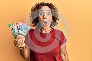 Beautiful middle age mature woman holding australian dollars scared and amazed with open mouth for surprise, disbelief face